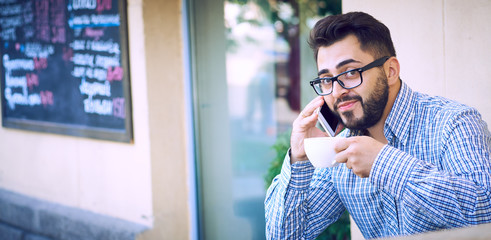 Modern hipster businessman with beard and glasses drinking coffee in the city cafe during lunch time and using mobile phone.