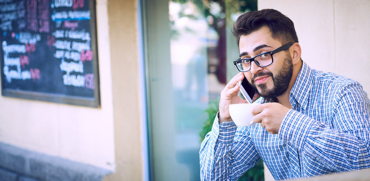 Modern hipster businessman with beard and glasses drinking coffee in the city cafe during lunch time and using mobile phone.