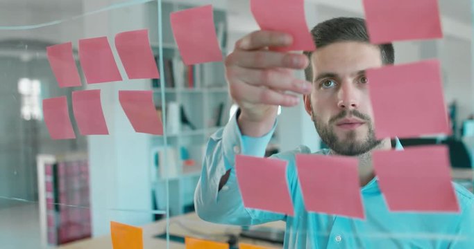 Caucasian male worker standing near glass wall with sticky notes, framework for managing work, scrum methodology. 4K UHD 60 FPS SLOW MOTION