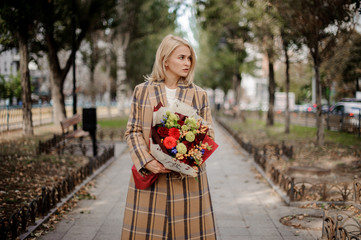 Blonde woman in plaid coat holding a bright bouquet of flowers standing on alley