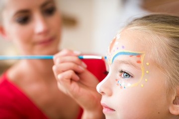 Young mother painting daughters face for Halloween party. Halloween or carnival family lifestyle background. Face painting and dressing up. - obrazy, fototapety, plakaty