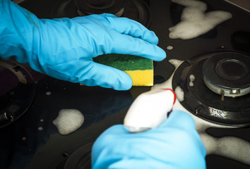 Human hand in a blue rubber glove washes the stove by gas detergent.