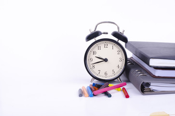 Stack of book with ticking vintage clock, artificial flower plant and colorful crayon on white table.View from above with copy space for text
