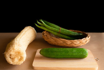 Luffa cylindrica (L.) M.J.Roem and Trichosanthes anguia Linn. Green fresh on chopping board with basket weave and dry sponge gourd. Black background and shadow. - obrazy, fototapety, plakaty