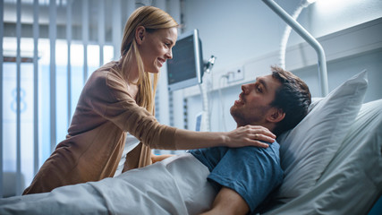 Obraz premium In the Hospital, Happy Wife Visits Her Recovering Husband who is Lying on the Bed. They Lovingly Embrace and Smile.