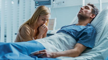 In the Hospital Sick Man Lying on the Bed, His Visiting Wife Hopefully Sits Beside Him and Prays...
