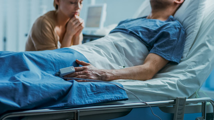 Obraz premium In the Hospital Sick Man Lying on the Bed, His Visiting Wife Hopefully Sits Beside Him and Prays for His Rapid Recovery. Tragic, Somber and Melancholy Scene.