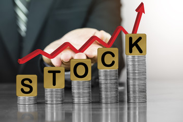 Businessman hand holding red arrow up with the letter stock on money coin stack arranged as a graph, concept of Increased investment and stock exchange