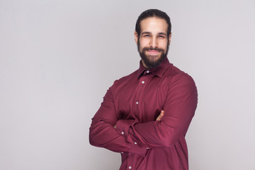 Portrait of handsome businessman with dark hair and beard in red shirt standing with crossed hands, looking at camera with satisfied face and smiling. indoor studio shot, isolated on gray background.