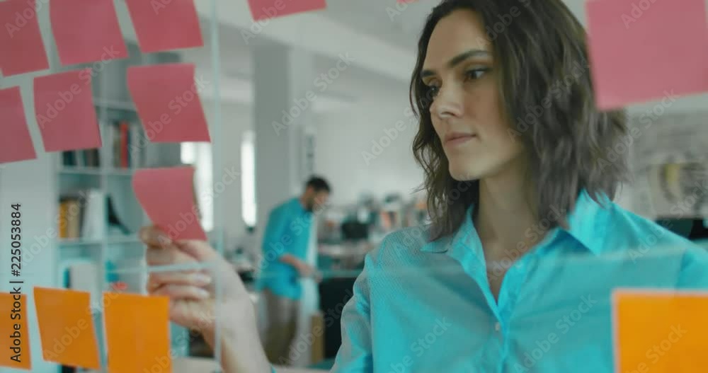 Poster Caucasian female worker standing near glass wall with sticky notes, framework for managing work, scrum methodology. 4K UHD 60 FPS SLOW MOTION