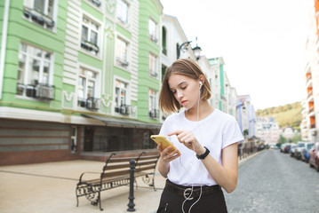 Concentrated attractive girl in a white T-shirt, uses a smartphone and listens to music in headphones at the background of the old street. Hipsters girl walking around the town