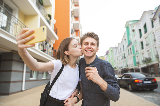 Portrait of a happy young couple: girl kisses her boyfriend in a cheek and takes a selfie on a smartphone, young man smiles with a cup of coffee in her hands.Couple take selfie background of the town