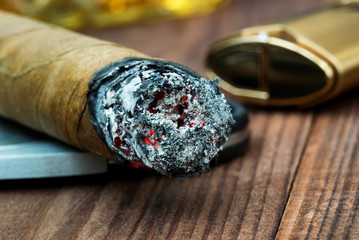 Smoldering cigar with ash on the end close-up next to a lighter and a piece of a glass of whiskey