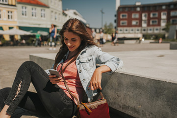 Fototapeta premium Young beautiful woman sitting in the city center holding a smartphone, smiling.