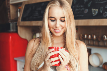 Nice blonde long hair girl wearing sleapwear standing in the red loft comfortable sunny kitchen, warming one's hands of the cup of coffee and enjoying the rays of the sun