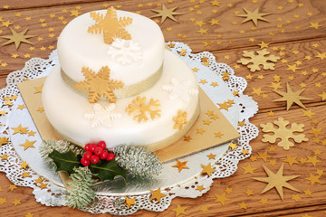Luxury Christmas cake with two tiered iced layers with snowflake and gold star decorations, winter holly and fir on oak wood background.