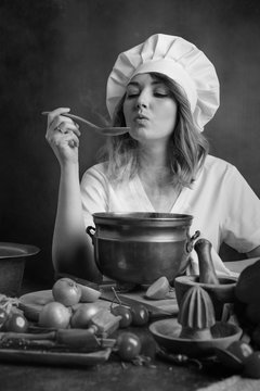  Young beautiful girl in a chef uniform with old pan and wooden spoon tasting food.