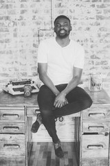 Attractive bearded African man sitting on the wooden table at his modern home studio. Concept of young people enjoying mobile devices. Brick wall on blurred background. black and white portrait