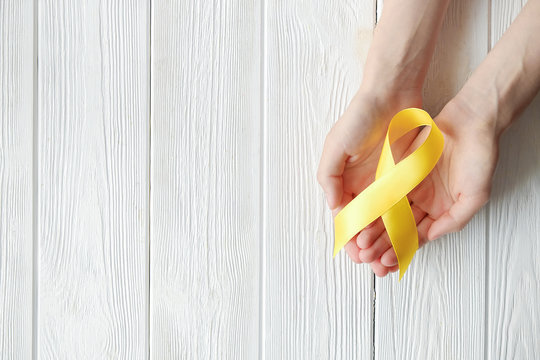 Woman's hand holding yellow ribbon - bladder, liver and bone cancer awareness symbol. Children cancer prevention concept. Isolated background, copy space, close up, top view - Powered by Adobe