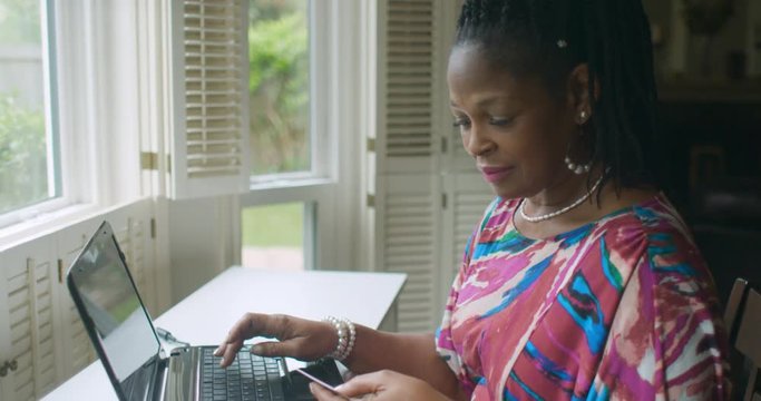 A lovely African American lady at home with laptop and credit card in hand sits by a large window enjoying a little sunshine as she shops online.
