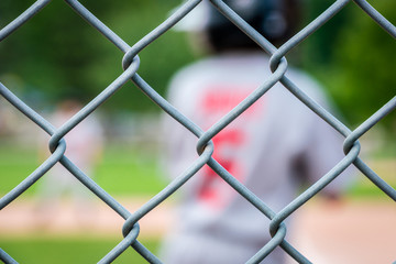 Unidentifiable youth junior league baseball player in team uniform, seen from behind through wire fence.