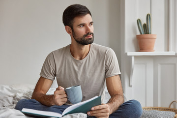 Indoor shot of pensive young Caucasian guy with thick beard, being deep in thoughts after reading...