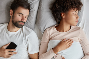 Young interracial family couple sleeps in bed, hold contemporary mobile phones, being addicted to...