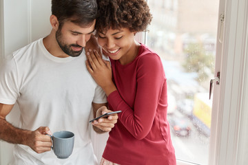 Lovely African American woman and her boyfriend watch video online via cell phone, enjoy togetherness, spend weekend at home during rainy weather outside, drink hot beverage, stand near window