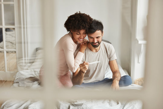Happy mixed race future parents look joyfully at pregnancy test, rejoice positive news about pregnancy, sit together on bed against domestic interior. Newlywed couple wait for new member in family
