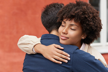 Carying woman embraces her boyfriend, has pleased facical expression, Afro haircut, pose against...