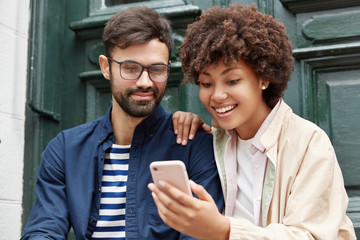 Attractive hipster and his dark skinned girlfriend watch funny video online, make shopping, use...