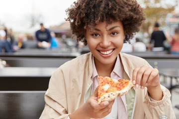 Photo of satisfied teenage girl with dark healthy skin, enjoys delicious meal, holds piece of...