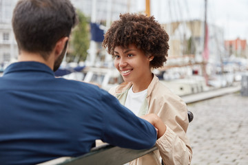 Horizontal shot of romantic couple in love sit on bench against sea port background, have lovely talk, discuss fututre plans, enjoys calm atmosphere, fresh marine breath. People and rest concept