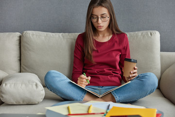 Horizontal shot of attractive brunette young woman writes plan for project, sits crossed legs on sofa, tries to concentrate on work, drinks takeaway coffee, wears casual red sweater and jeans.