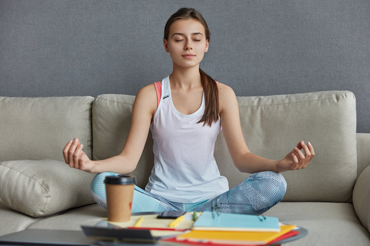 Relaxed Concentrated Teenager Dressed In Casual Clothes, Sits In Yoga Pose, Meditates And Has Break After Hard Work, Drinks Takeaway Coffee, Poses On Comfortable Couch At Home. Meditation Concept