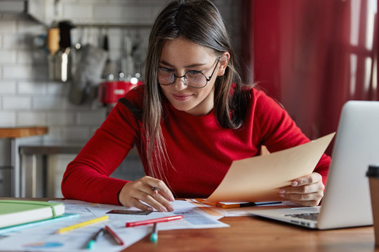Horizontal shot of cheerful woman sits at kitchen table reviews finances, sits in front of opened laptop, wears red turtleneck sweater, makes notes in papers, cuts off expenses. Domestic interior