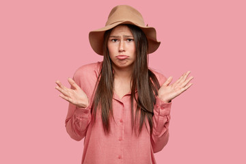 Isolated shot of displeased brunette woman purses lips, cant make right decision, feels insulted, dressed in fashionable outfit, expresses apathy, poses against pink background. Puzzlement concept