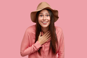 Pleased happy girl keeps hand on chest, smiles broadly, cant believe in unexpected surprise, being...