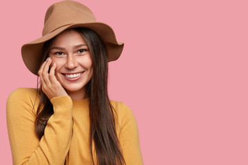 Indoor shot of attractive young woman with dark hair, smiles positively at camera, keeps hand on...