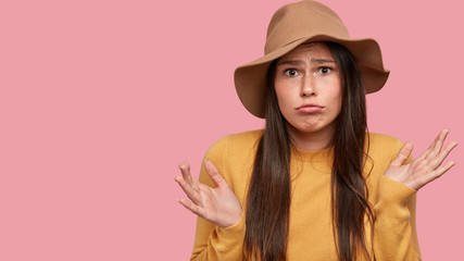 Uncertain displeased woman with dark long hair, freckled skin, shrugs shoulders with hesitation, cant make choice between two suggestions, poses against pink background, free space for you on left