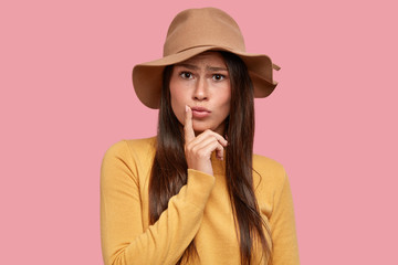 Displeased indignant teenager pouts lips, keeps finger near mouth, wears fashionable hat, casual...