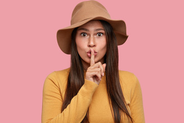 Hush, please! Beautiful young brunette woman keeps index finger over lips, has surprised...