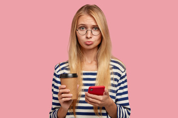 Sad blonde woman with offensive expression, purses lips in displeasure as recieves bad message from...
