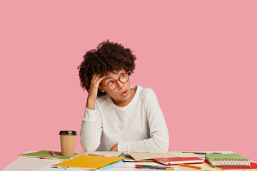 Shocked African American woman wears glasses, dressed in white clothes, works on creation startup, poses against pink background. Wondered dark skinned young entrepreneur stunned with much work