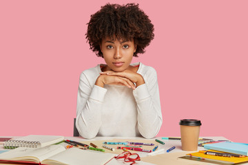 Indoor shot of serious African American architect keeps hands under chin, looks direclty at camera, dressed in white jacket, uses colourful markers and notebook for making sketches, poses indoor