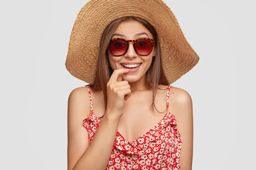 Studio shot of attractive woman has summer vacation, smiles positively and keeps finger near mouth, dressed in fasionable clothes, trendy sunglasses, poses over white background. Leisure concept