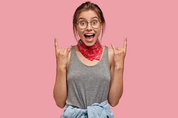 Fashionable hipster woman makes rock and roll gesture, excalims happily being in high spirit,...