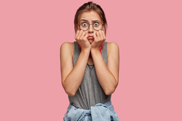 Terrified trembling woman stares in stupor with eyes popped out, expresses fear and shock, wears casual outfit and round spectacles, poses against pink background. Nervous girl poses indoor.