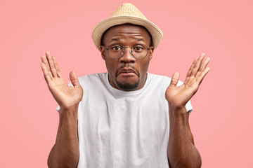 Isolated shot of clueless dark skinned man with unaware expression, shrugs shoulders, expresses apathy, dressed in casual t shirt, straw hat, models against pink background. Hesitation, bewilderment