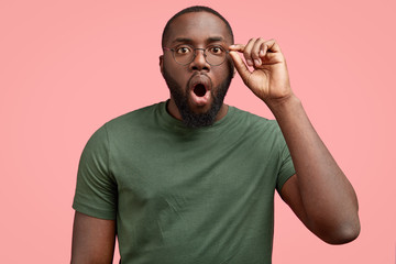 Over emotive dark skinned shocked unshaven African American man stares through spectacles, keeps...